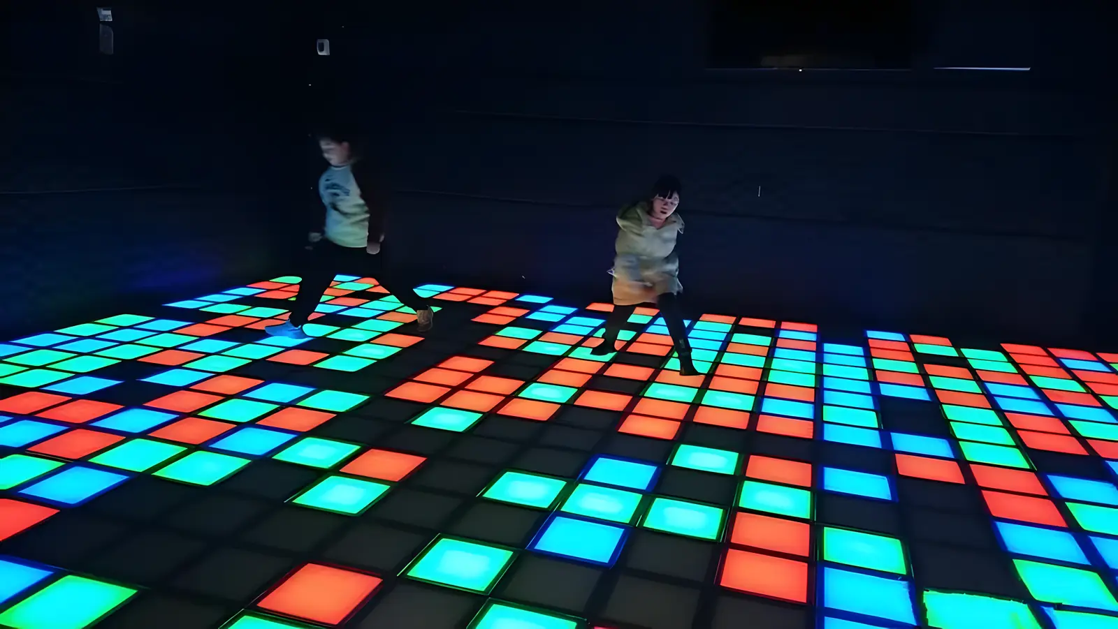 Two kids jumping across a floor of glowing red, green and blue tiles